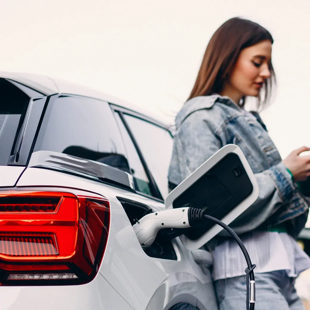 Woman charging an electric car, close-up of the vehicle's rear lights and charging station on one side. The background is blurred. She has long brown hair