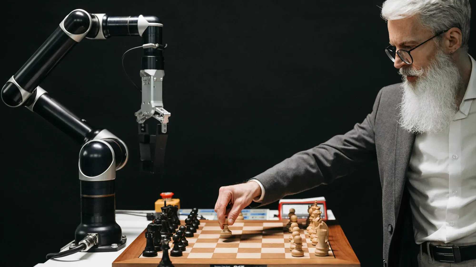 A man with a white beard and glasses playing chess against an AI robot. The setting is on a black background, with a single robotic arm that plays for him.