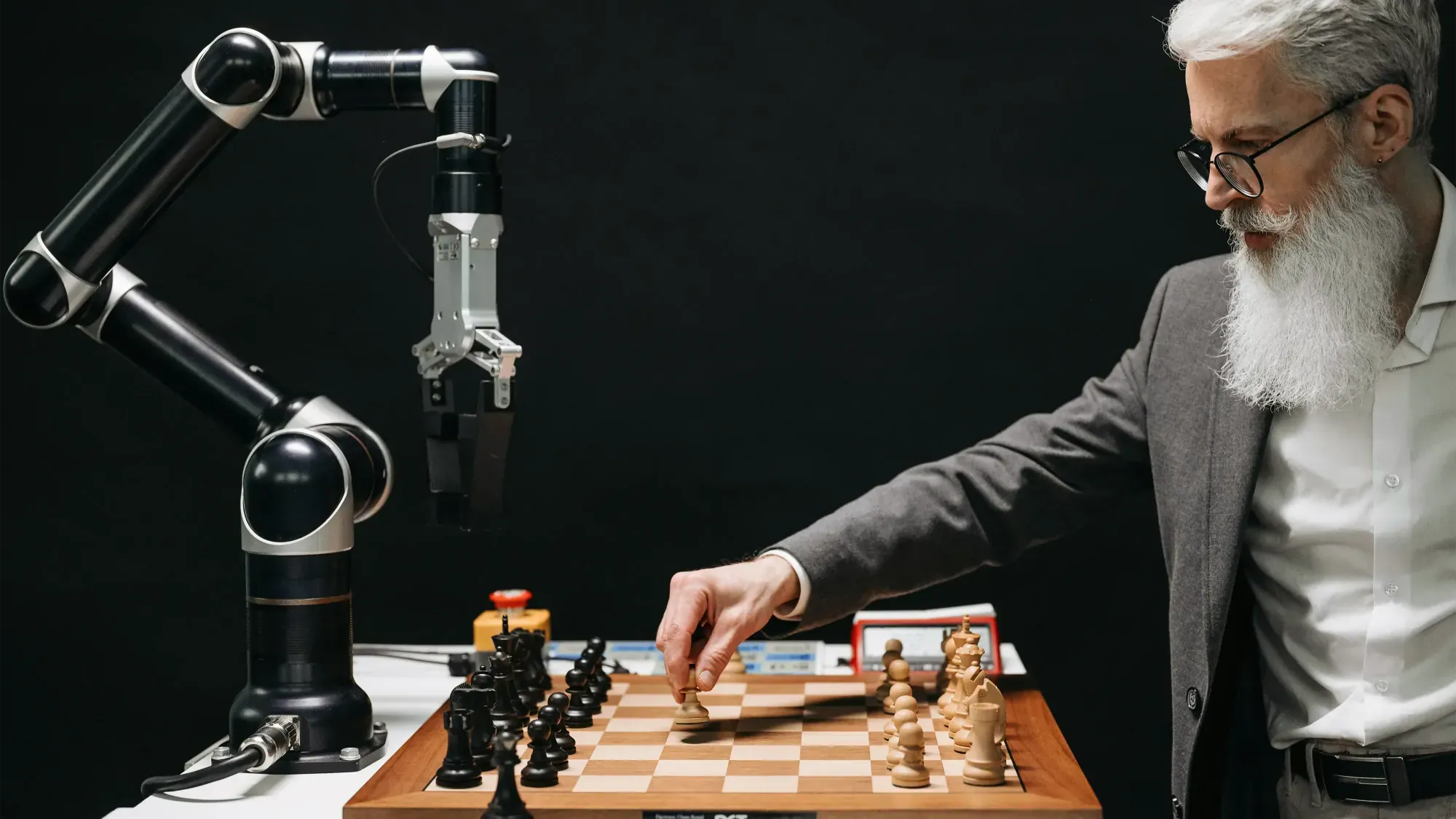 A man with a white beard and glasses playing chess against an AI robot. The setting is on a black background, with a single robotic arm that plays for him.
