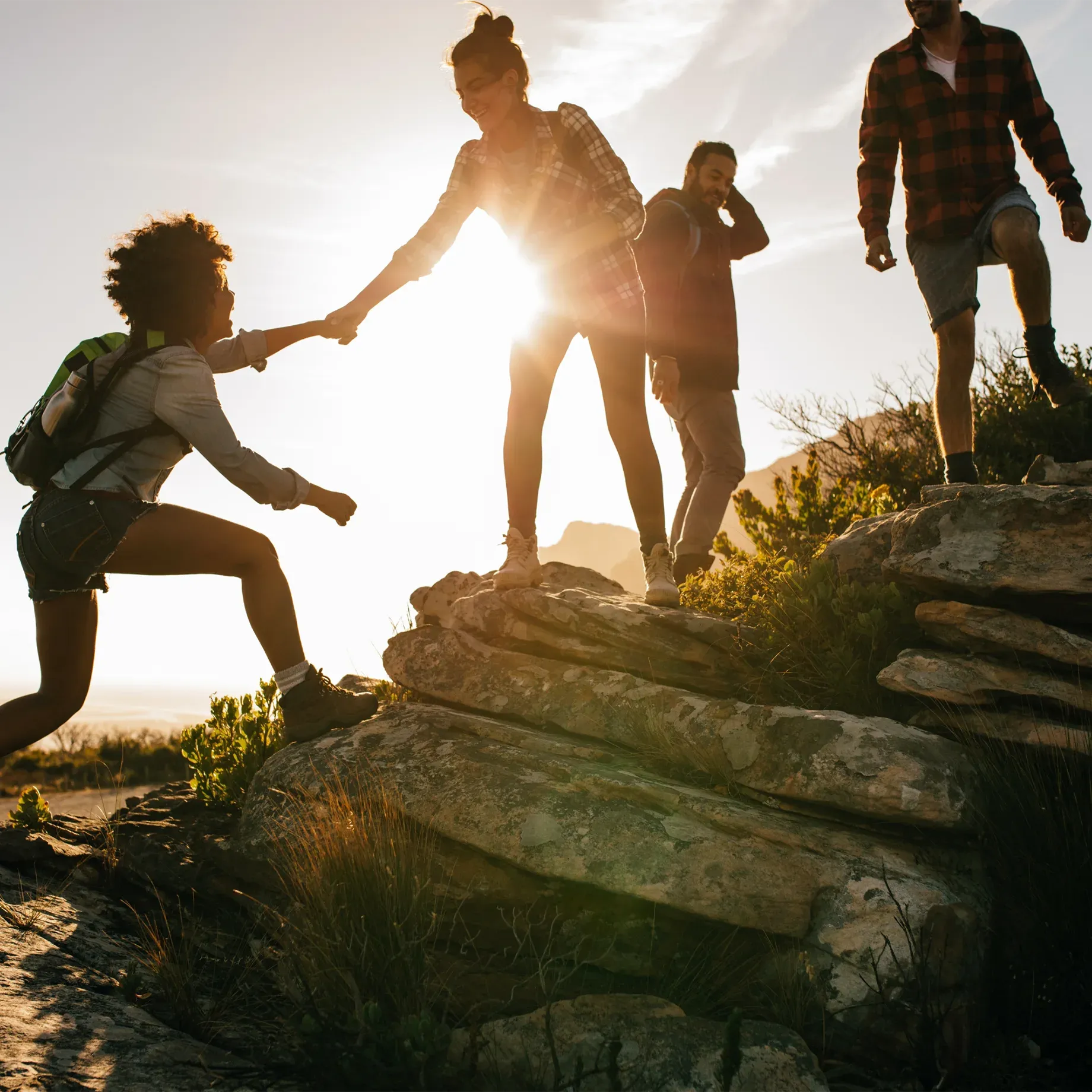 A group of friends hiking on rocky terrain, one helping the other up over rocks with their hands outstretched towards each other. The sun is setting in the background, casting long shadows and highlighting them all against the sky.