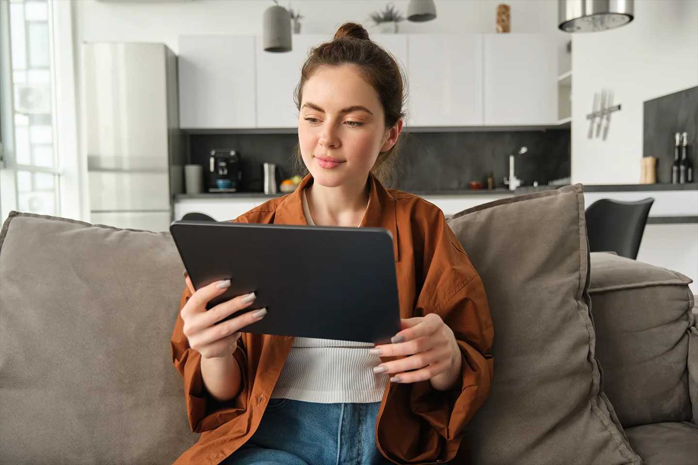A young woman sits on the couch in her modern apartment, using a tablet computer.