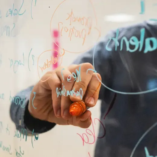 A close-up of an open hand writing on a glass with a marker, with the focus on the tip and pointed finger of the right hand.