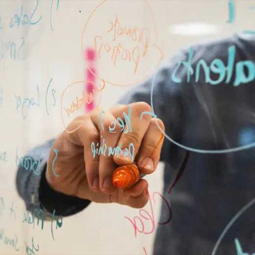 A close-up of an open hand writing on a glass with a marker, with the focus on the tip and pointed finger of the right hand.