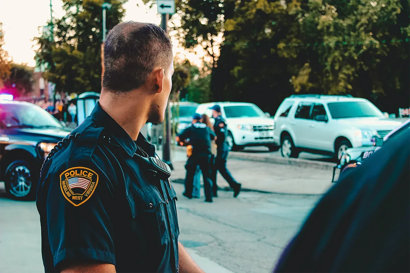 A police officer in uniform, speaking to other officers near parked cars and streetlights on the side of an urban road during daylight.