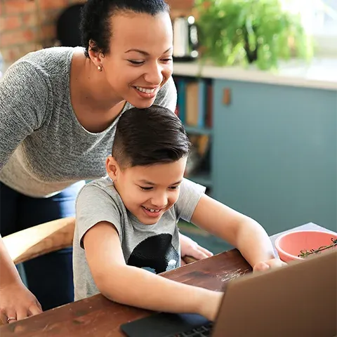 A young boy is sitting at the kitchen table, smiling and watching an online video on his laptop while being watched by his mother in a grey t-shirt.