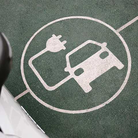 Top-down view of an electric vehicle charging bay, with a white EV charging symbol of a car and a power plug painted on green asphalt, and the edge of a parked car.