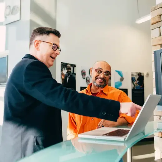 Office worker pointing at the screen on his laptop and smiling while talking to another man in front, both wearing glasses with black frames