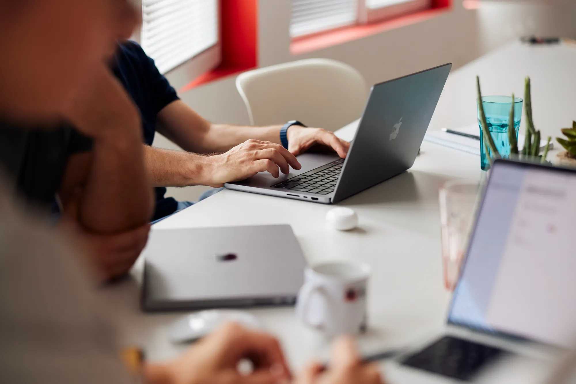 Engineers working on laptops around a shared table in an office with red window frames.