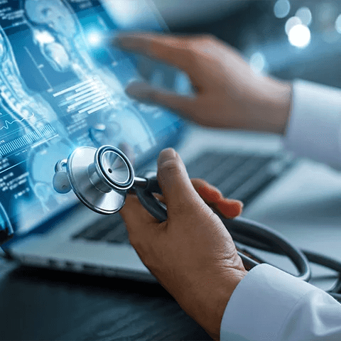Close-up of a doctor's hand holding a stethoscope. In the background, an open laptop displays medical images and data on the screen.