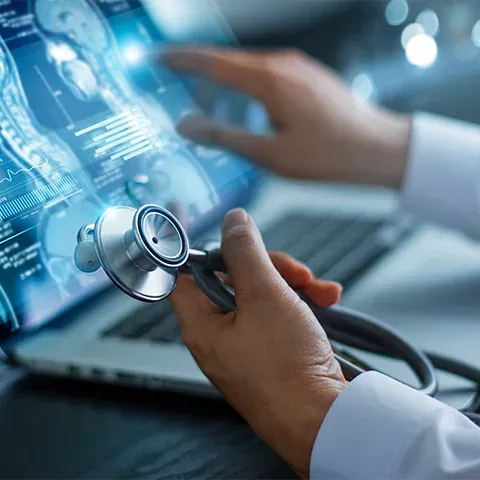 Close-up of a doctor's hand holding a stethoscope. In the background, an open laptop displays medical images and data on the screen.