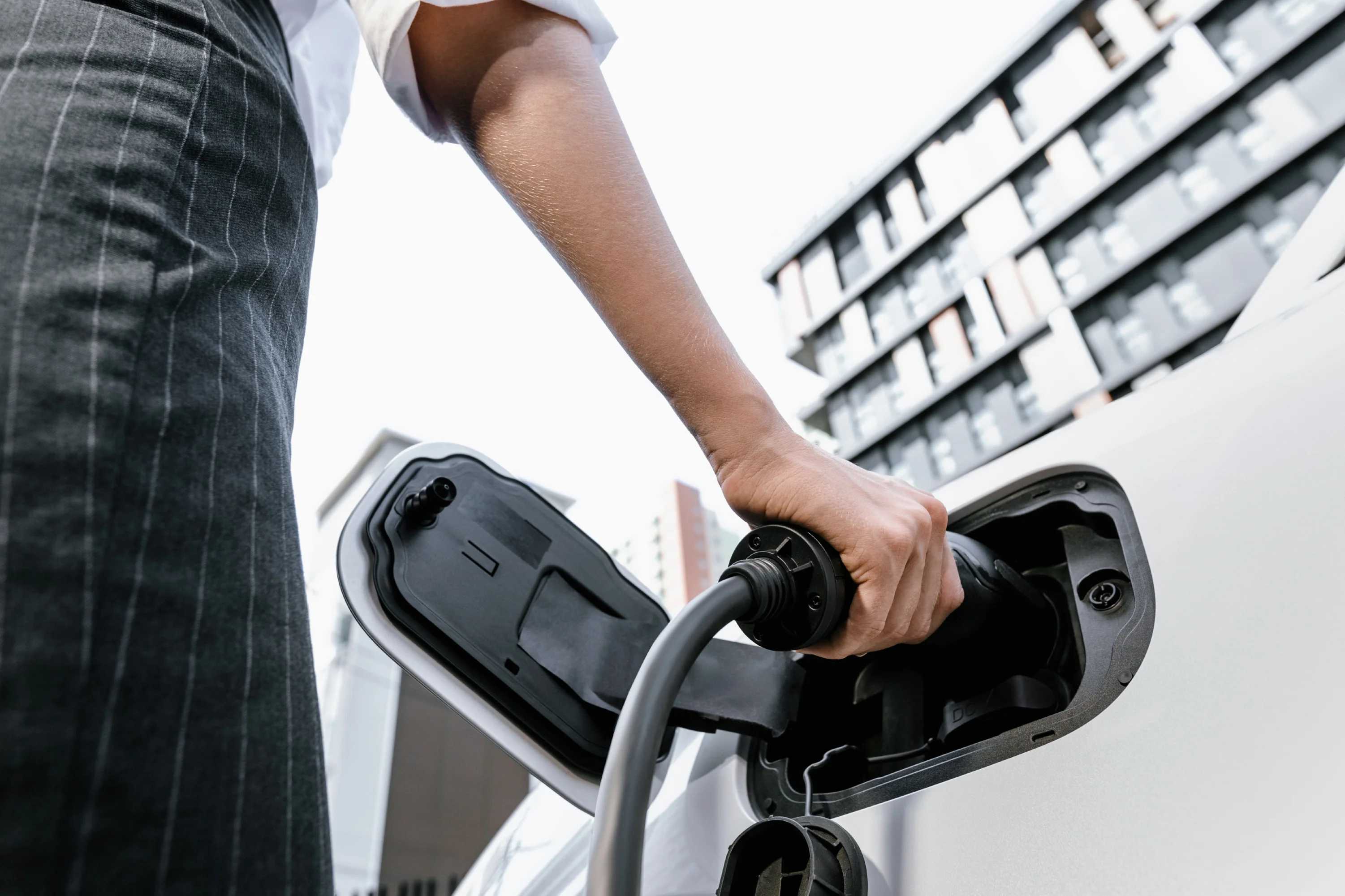 A driver plugging a connector into an EV fast charger in front of an office building.