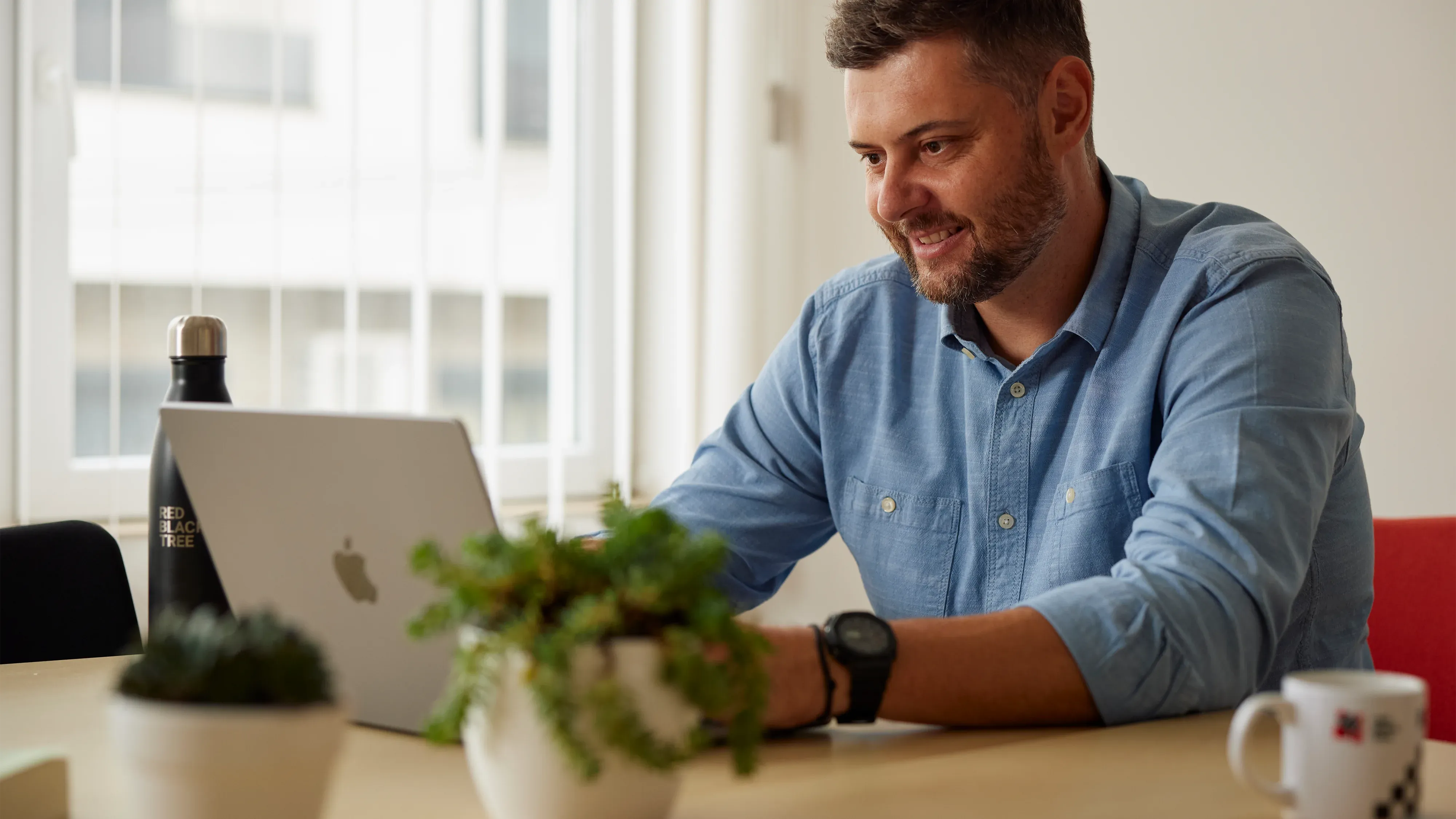 Man is sitting at the table and smiling while working on a MacBook Pro inside a modern office with white walls and red accents.