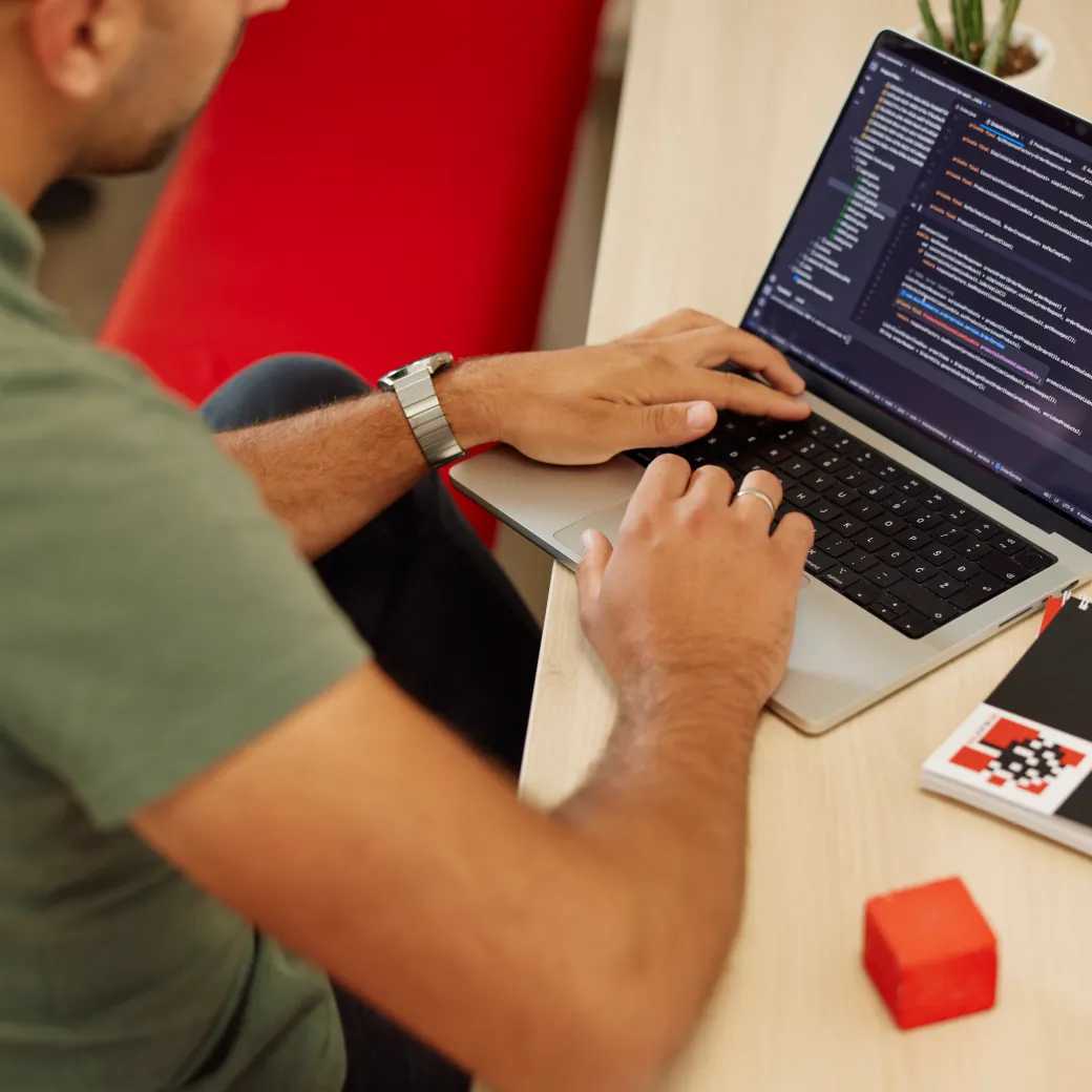 An engineer writing code on a laptop beside an RBT notebook and a red cube.