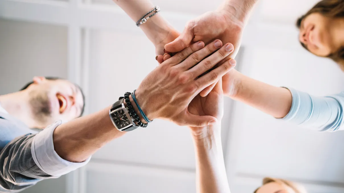 Close-up of five hands from different people stacked on top, showing unity and support in an office setting.