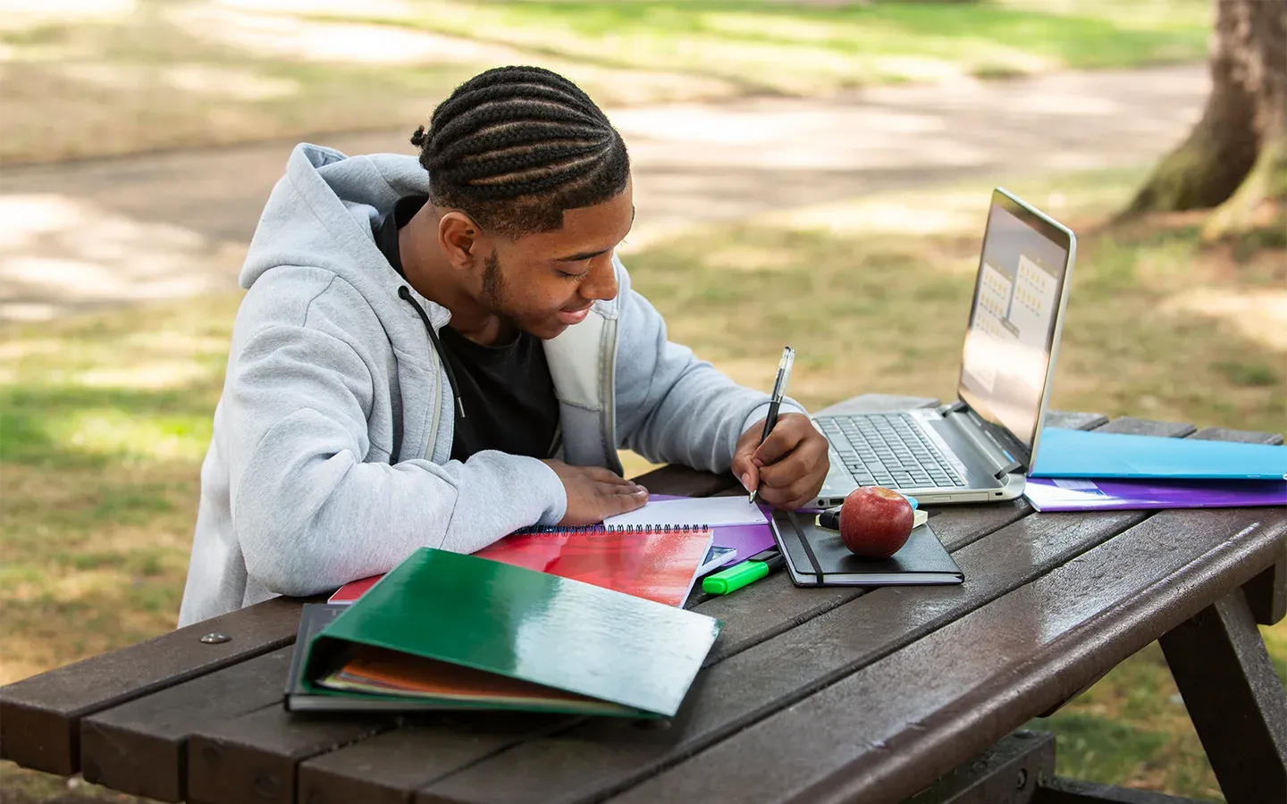 A male student is sitting at an outdoor picnic table writing in his notebook while using laptop.