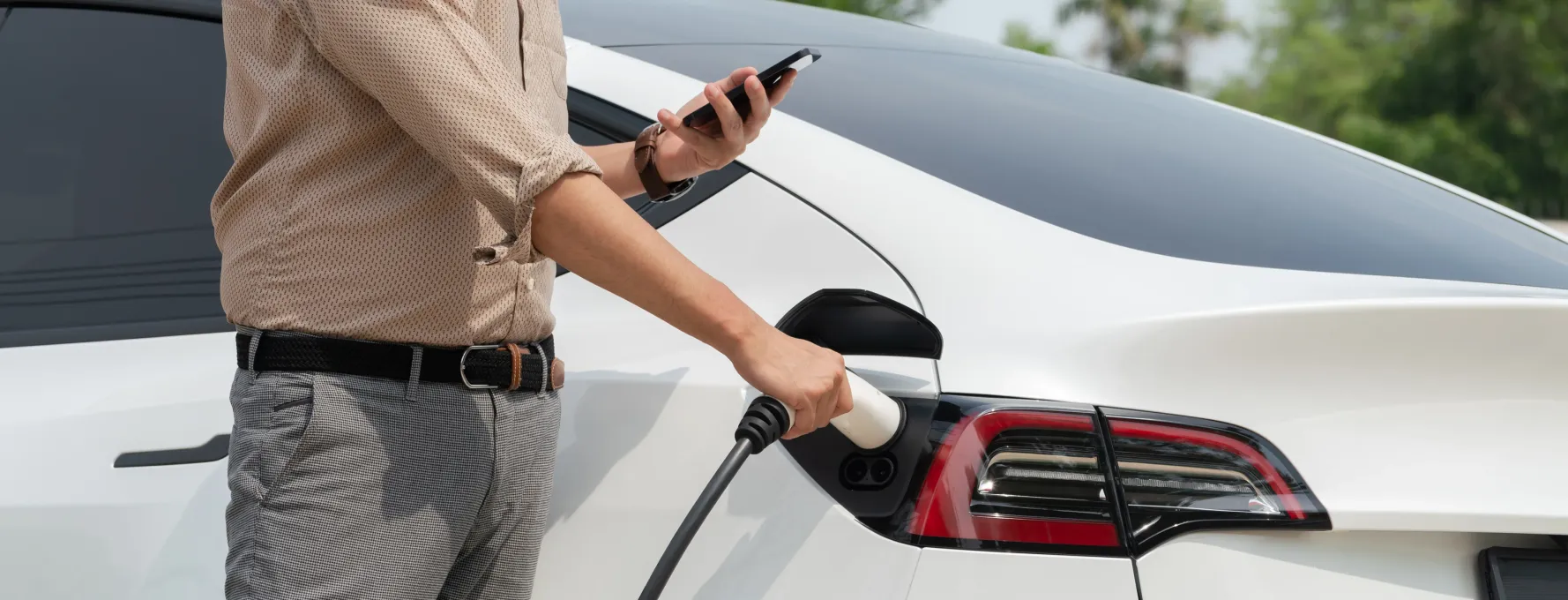 A man holding an electric car charging cable and looking at his phone.