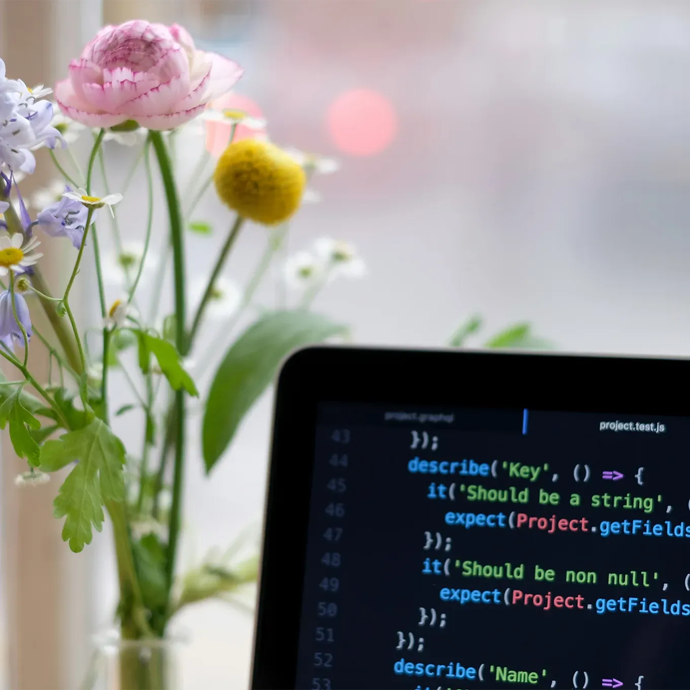 A close-up of an open laptop screen displaying lines of code. The background is blurred to show natural light coming through a window. A vase filled with flowers sits next to it.