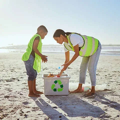 Photo of a mother and son wearing green and yellow reflective vests, picking up trash on the beach near their small white box with a recycling sign on it. It's a sunny day, and the beach appears clean.