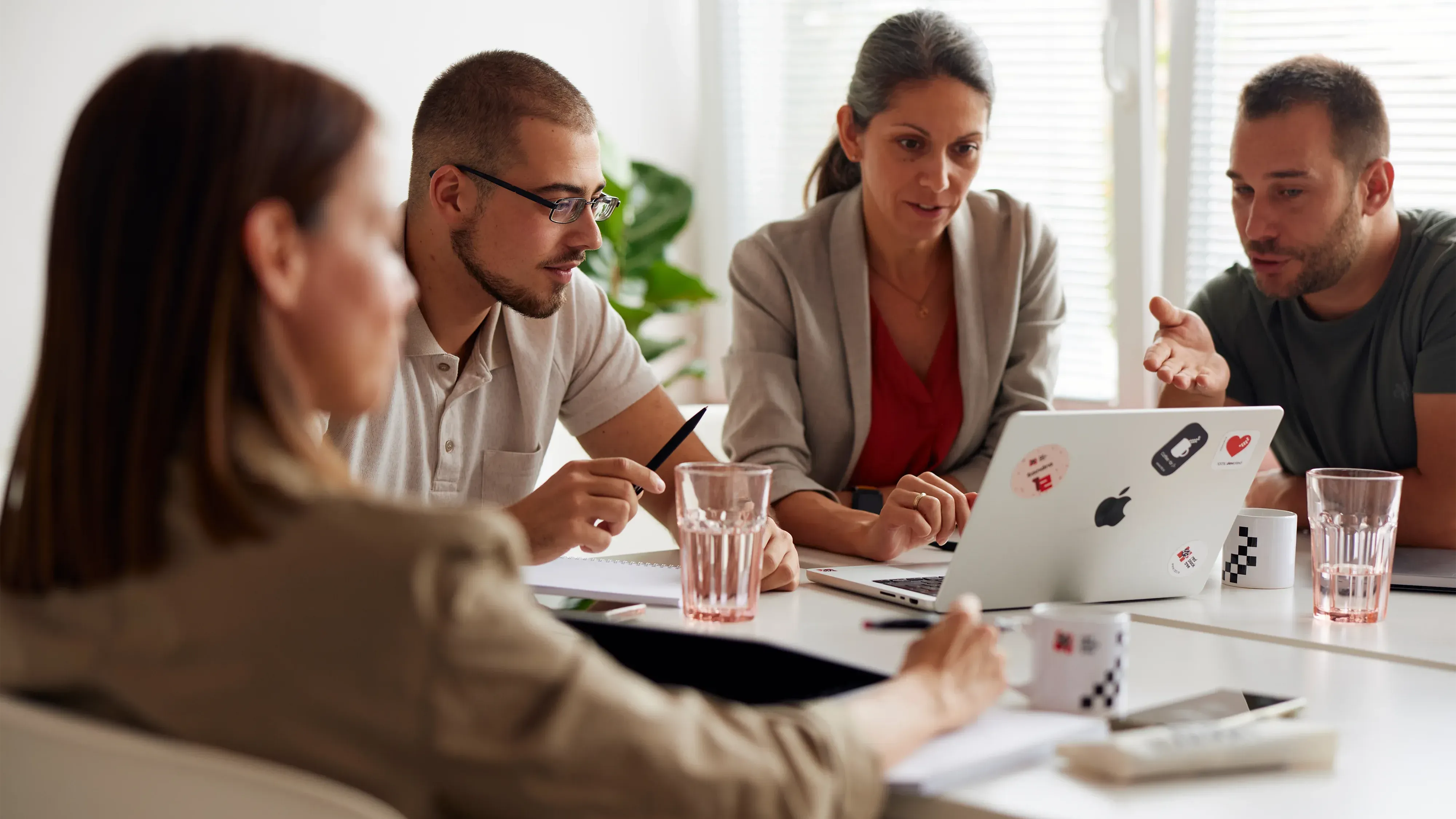 A group of people in an office meeting, sitting around the table with laptops and paper notes on it. They look focused as one man is pointing at his laptop screen. They are all dressed casually, in a bright white interior with soft lighting.