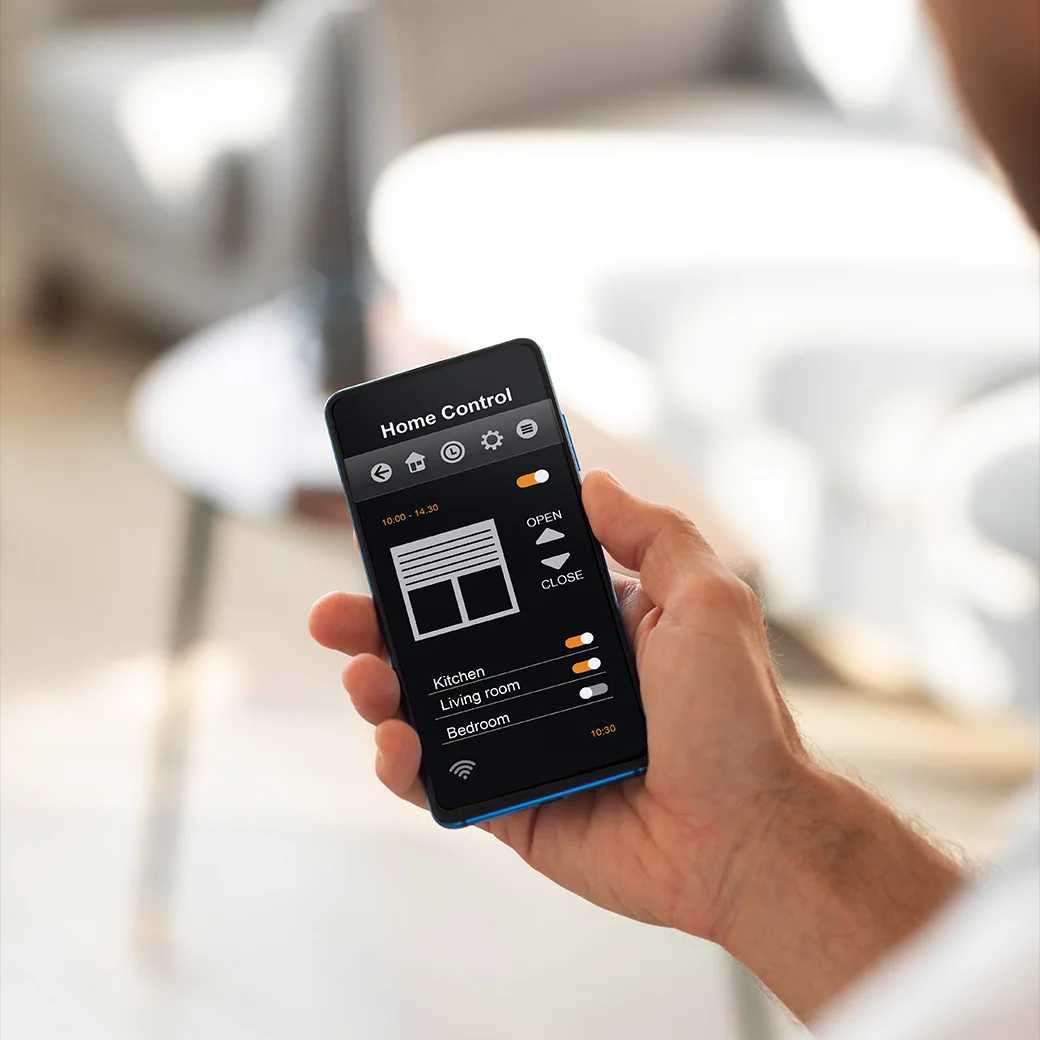 An over-the-shoulder shot of an individual holding and interacting with their phone, displaying the "Home Control" app interface on screen in a modern living room setting.