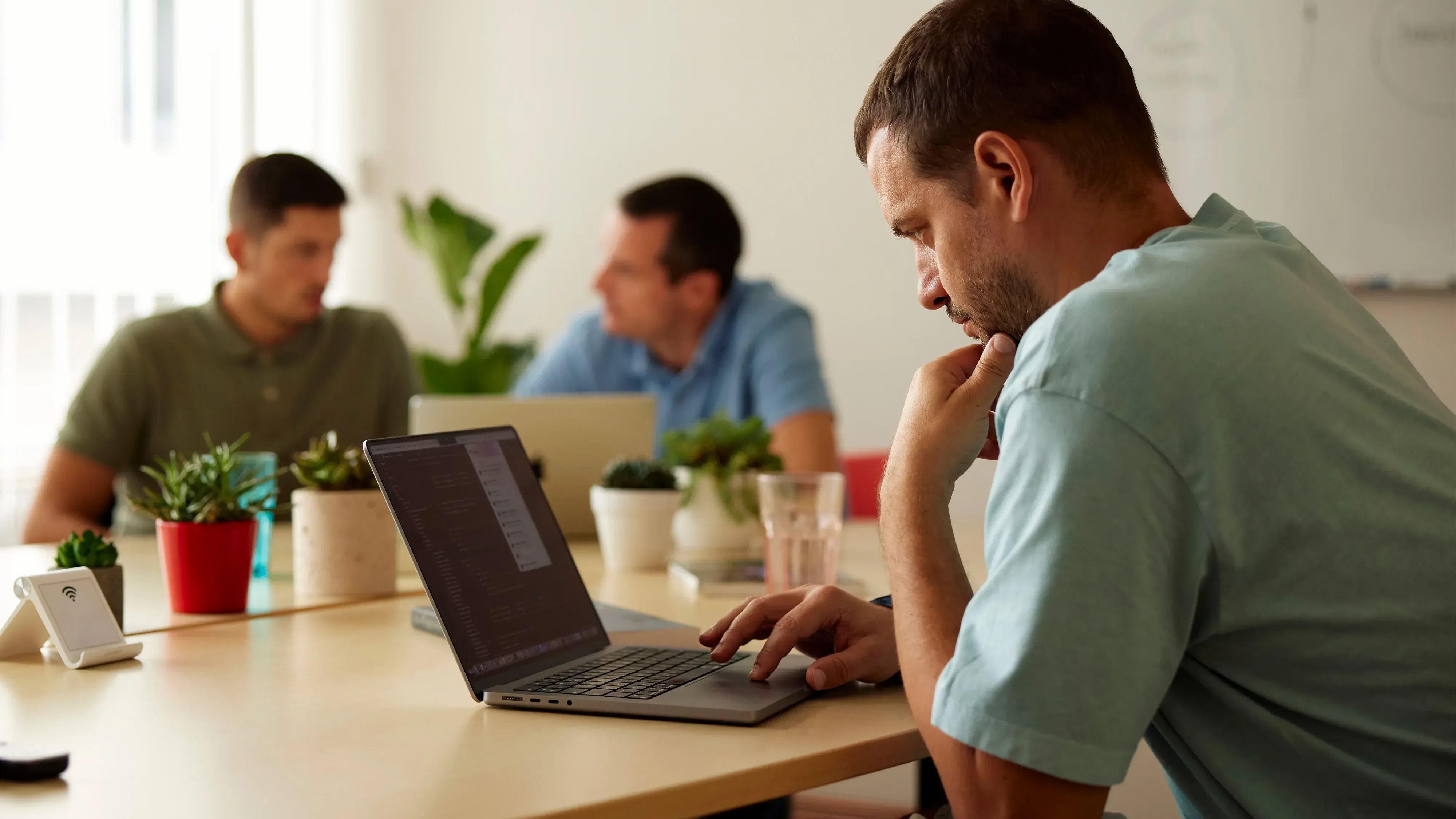 Three men sitting at a table, working on laptops in an office with white walls and green plants.