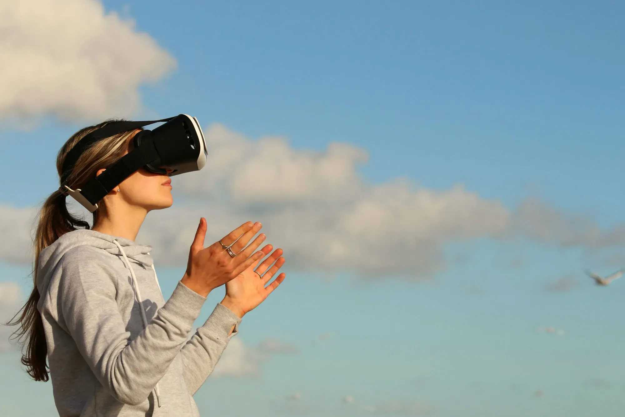 A woman wearing a VR headset outdoors, with her hands raised in the air, against a sky background.