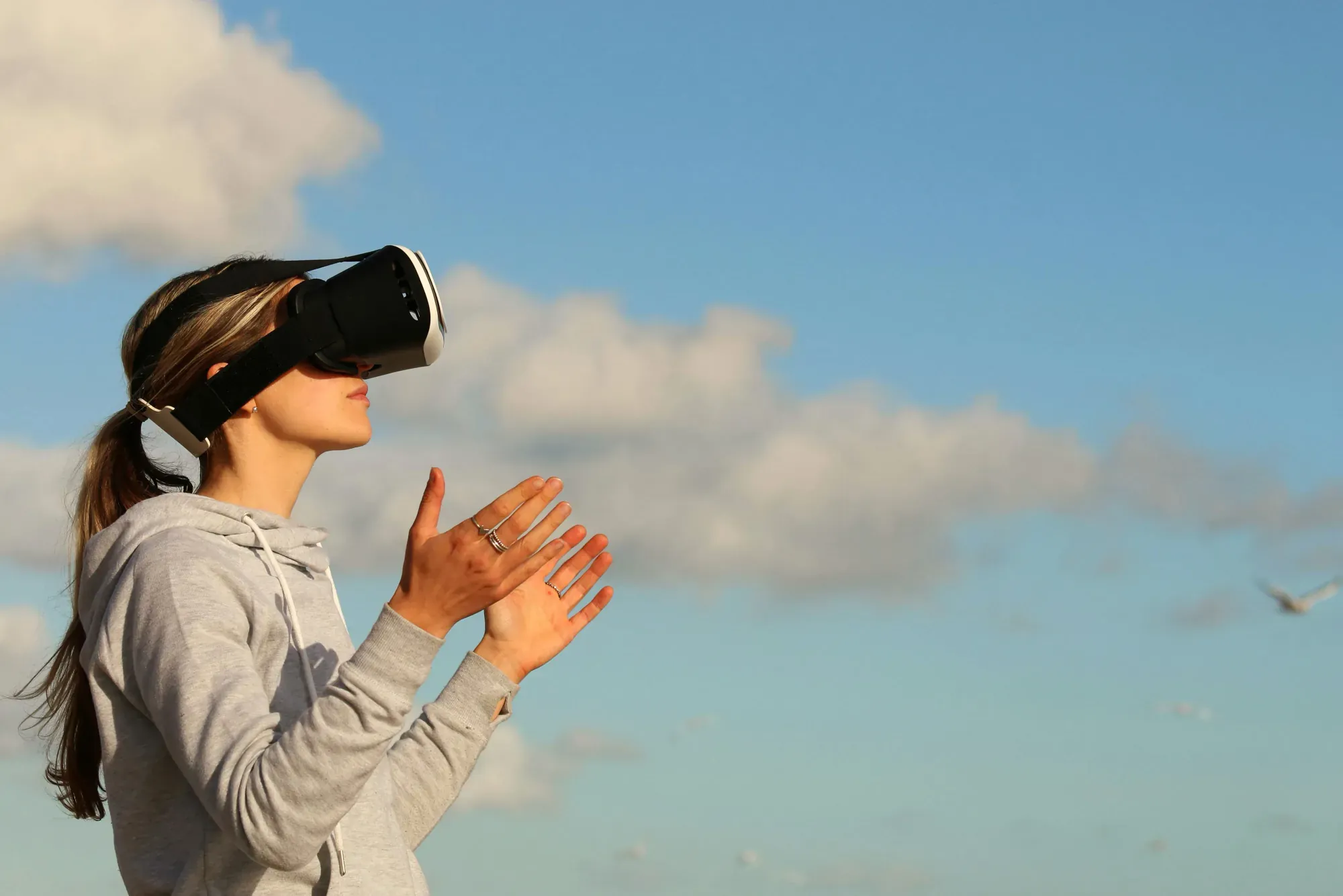 A woman wearing a VR headset outdoors, with her hands raised in the air, against a sky background.