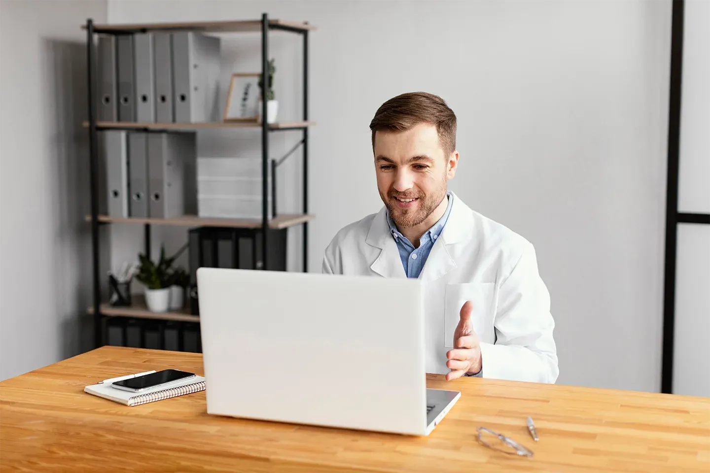 A male doctor, dressed in a white coat, sits at his desk, using a laptop for a video call with a patient.