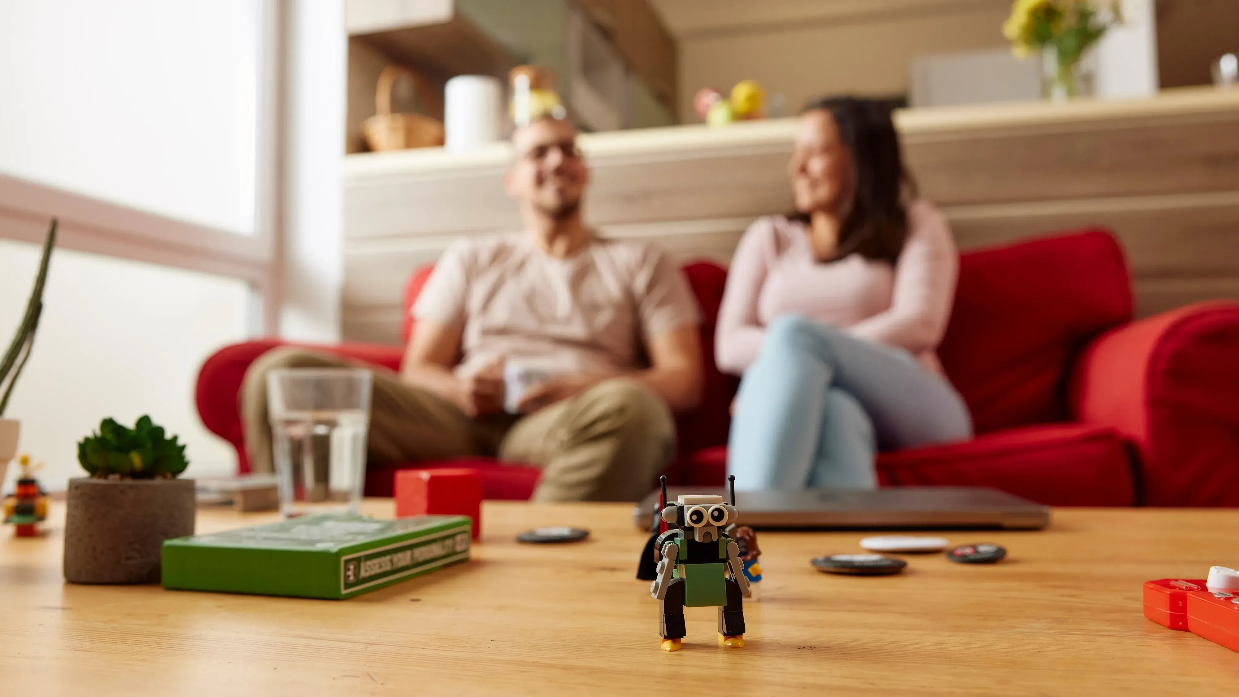 A small robot toy is on the table in front of two people sitting and laughing. The scene includes a sofa with red fabric, a wooden coffee table and a  room interior with light tones. The background is blurred, creating a depth of field effect.