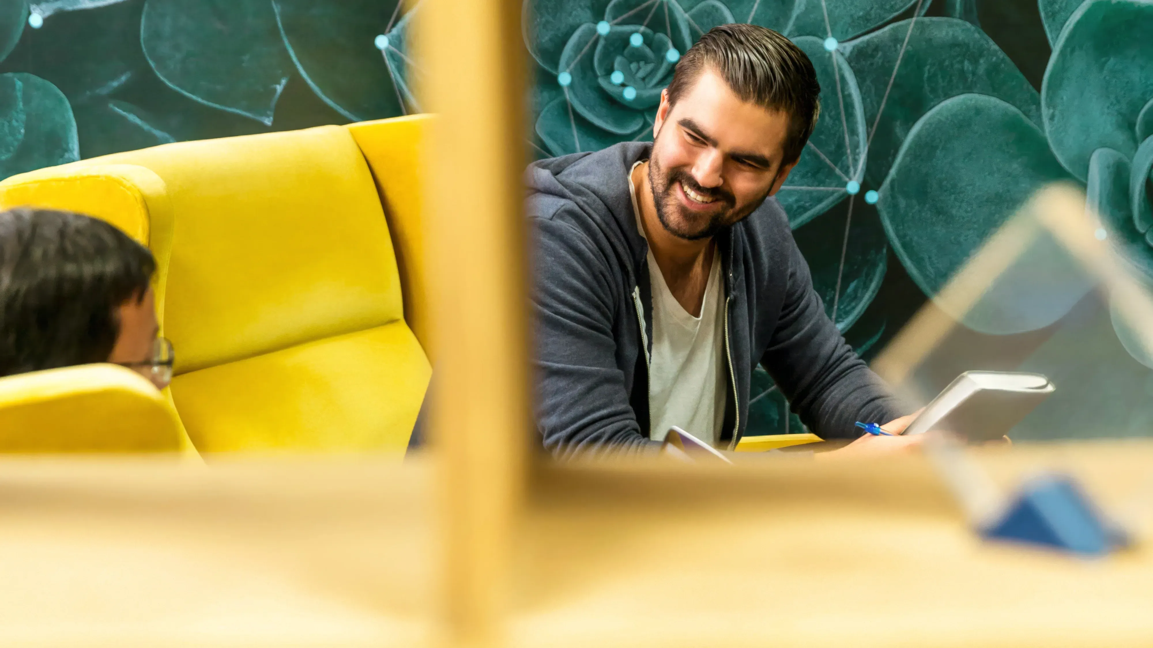 Two men smiling and laughing while working on laptops in an open space, surrounded by yellow sofas, with green plant patterns on the wall behind them.