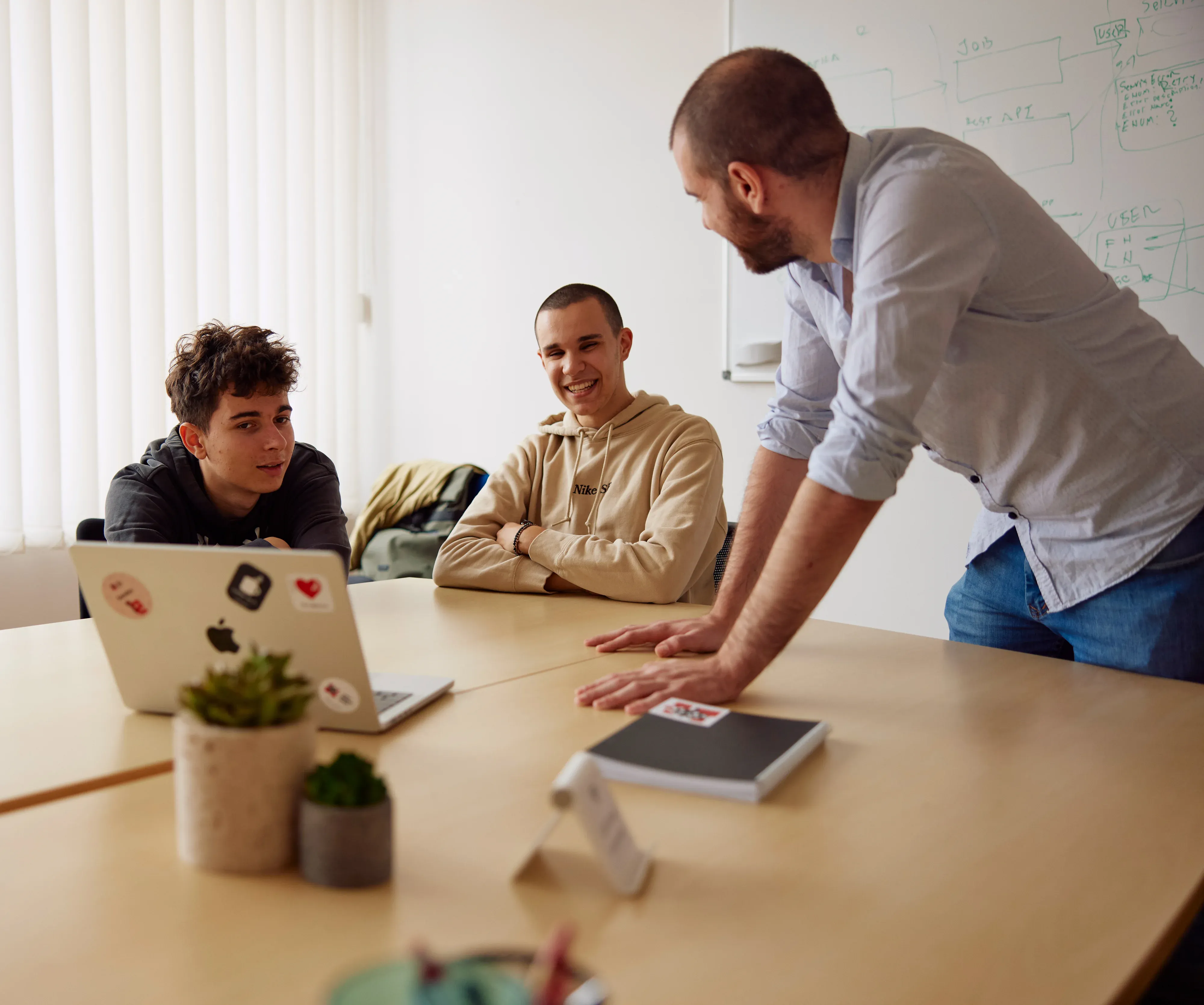 Two young men sitting at a table in an office, smiling and looking at a third man who is standing up with his hand on the desk near a laptop, with a whiteboard behind him.