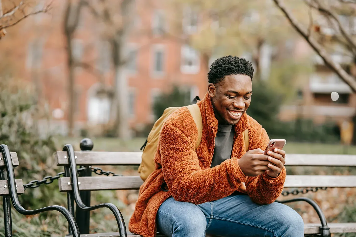 A young man is sitting on an outdoor bench, smiling and looking at his phone in the park.