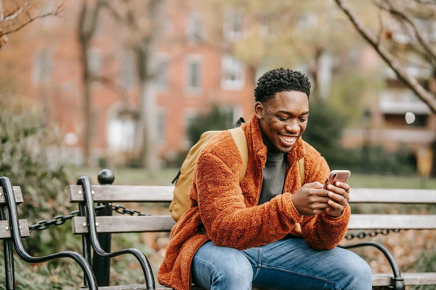 A young man is sitting on an outdoor bench, smiling and looking at his phone in the park.