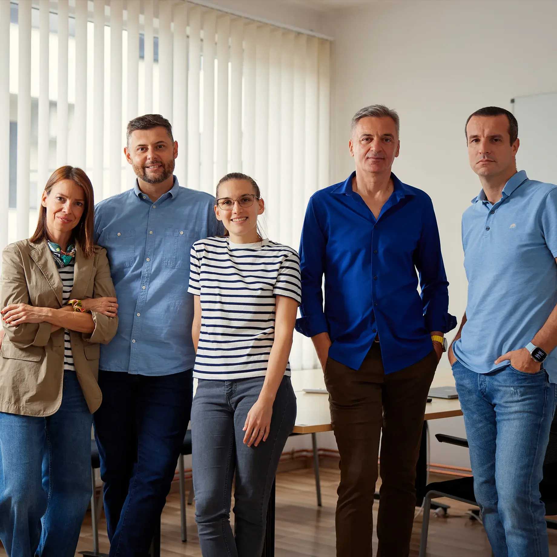 Five people standing in an office, three men and two women. The background features white vertical blinds, with natural light from a window on the left side.