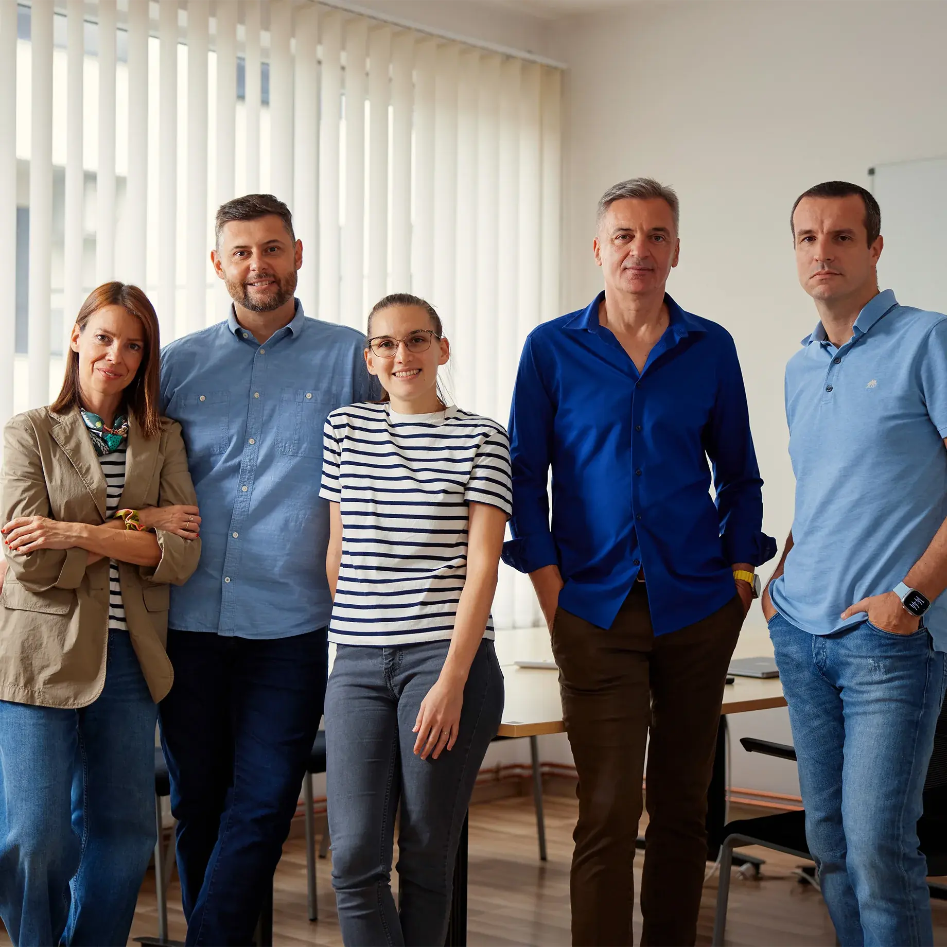 Five people standing in an office, three men and two women. The background features white vertical blinds, with natural light from a window on the left side.