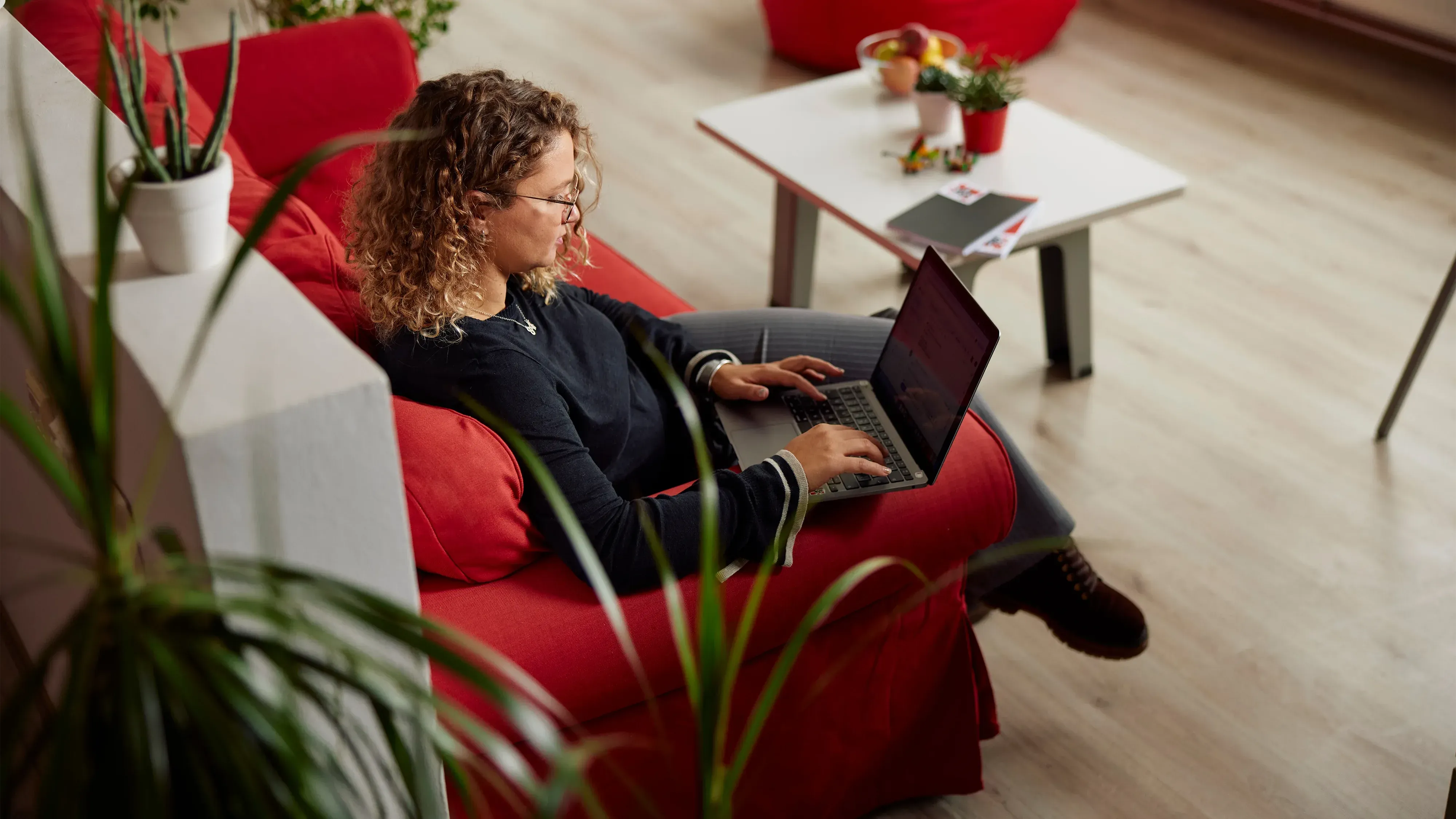 A woman sitting on a red couch in an open office space, working on her laptop computer.