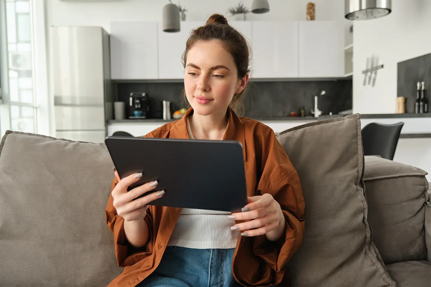 A young woman sits on the couch in her modern apartment, using a tablet computer.
